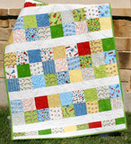 Handmade modern baby quilt featuring colorful novelty patchwork fabrics and charm pack quilt blocks displayed outdoors against a stone wall with detailed longarm quilting.